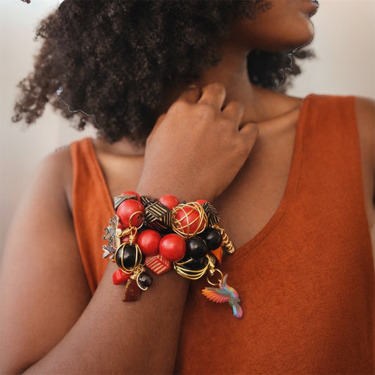 Brown skin woman with natural afro wearing chunky Afrocentric red and black bead charm bracelet stack with gold accents and hummingbird charm