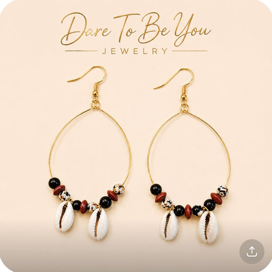Afrocentric cowrie shell gold wire hoop earrings with black beads, speckled beads, and wood accents on a cream background by Dare To Be You Jewelry.