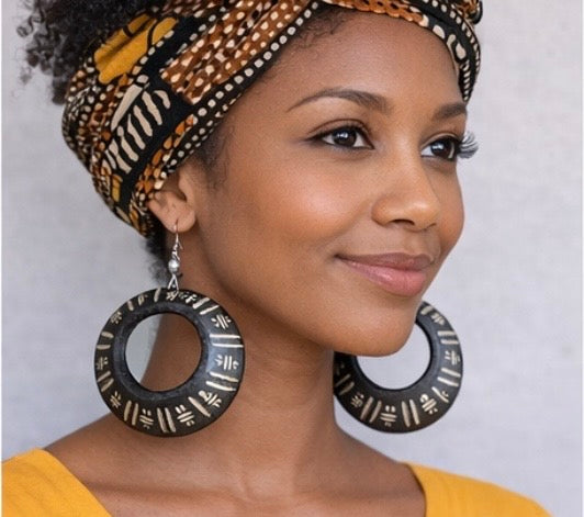 Brown skin woman wearing Afrocentric black hoop earrings with patterned headwrap and mustard yellow top