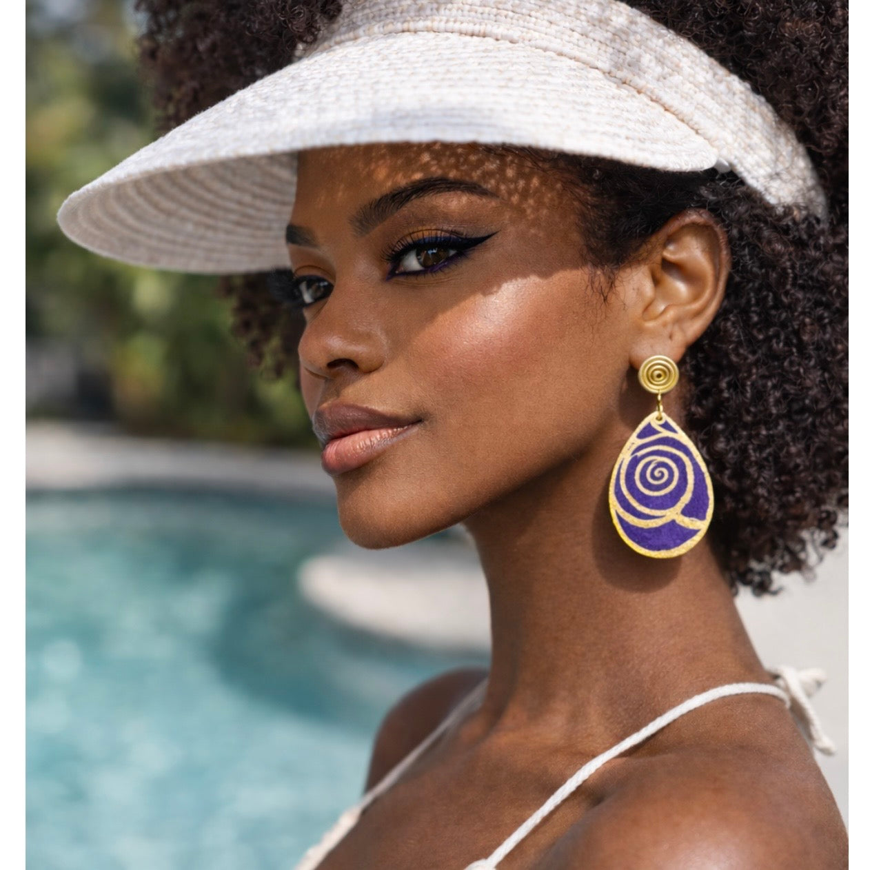 Dark skin woman wearing a sun visor and purple and gold Afrocentric statement earrings outdoors by a pool lifestyle image
