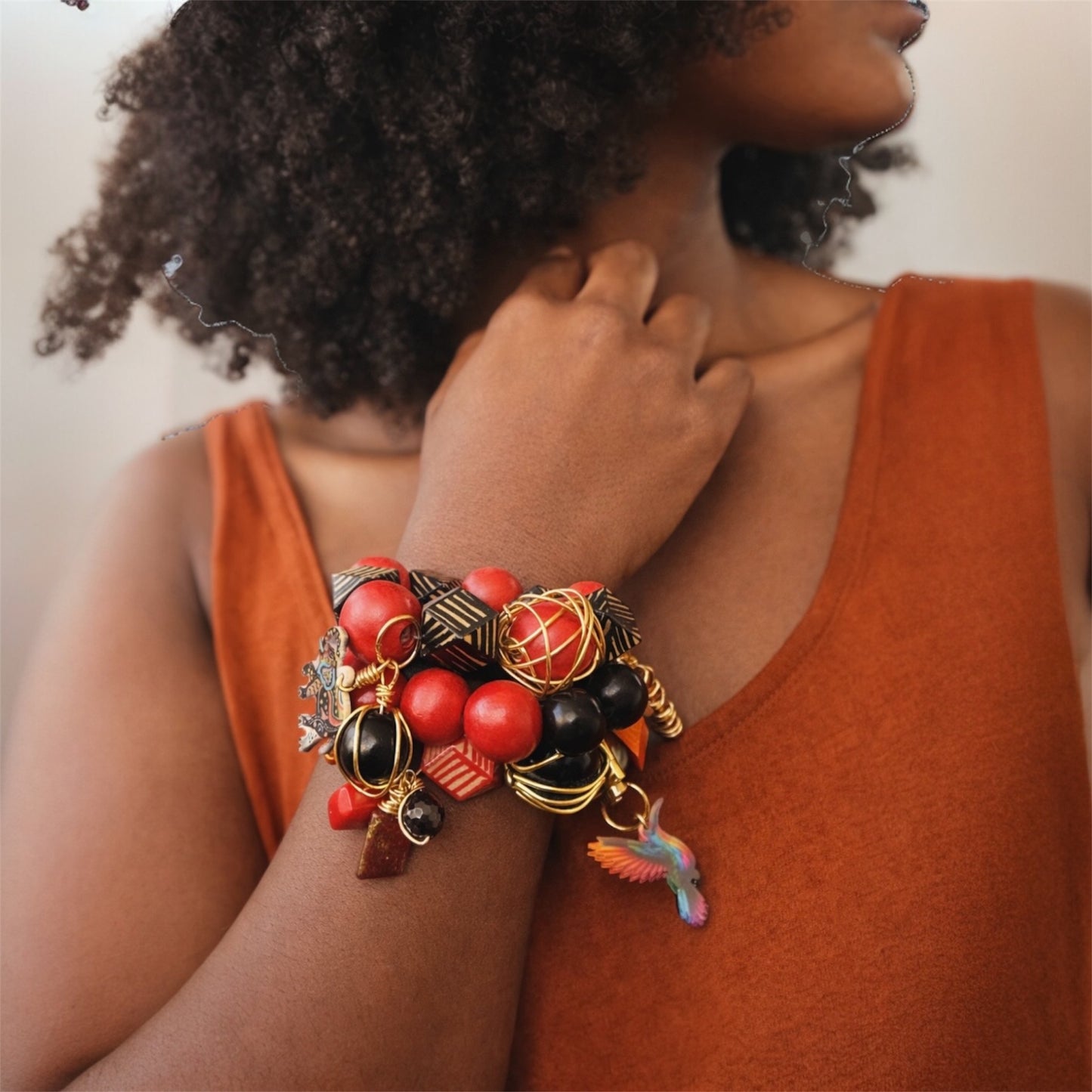 Brown skin woman with natural afro wearing chunky Afrocentric red and black bead charm bracelet stack with  gold accents and hummingbird charm