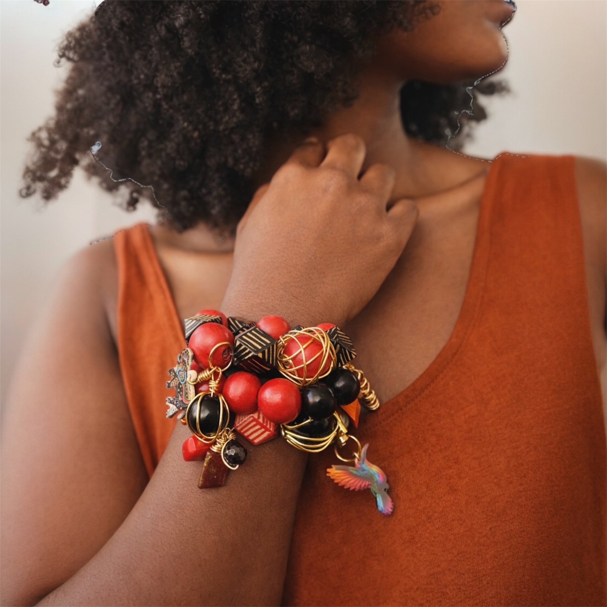 Brown skin woman with natural afro wearing chunky Afrocentric red and black bead charm bracelet stack with  gold accents and hummingbird charm