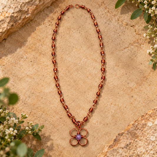 Handmade pure copper flower necklace featuring handcrafted wire-wrapped chain links and a lavender center bead, displayed on a warm earthy stone background with delicate greenery.