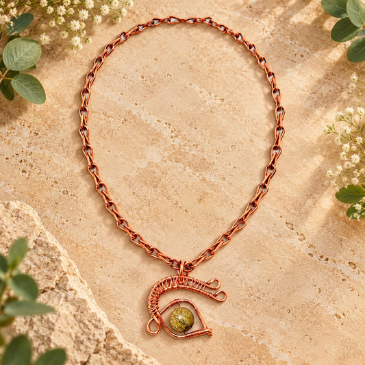 Handmade pure copper protective eye necklace featuring a wire-wrapped serpentine gemstone pendant and handcrafted copper link chain, displayed on a warm earthy stone background with greenery.